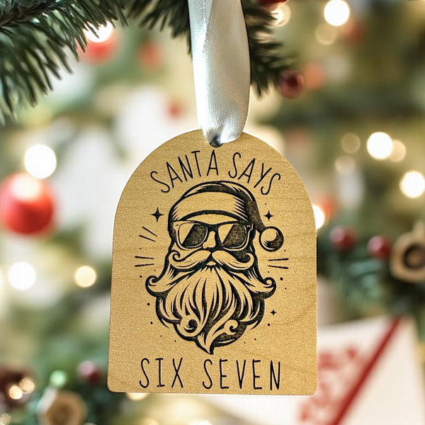 Christmas ornament tag with Santa Claus design and text 'Santa Says Six Seven' on a blurred Christmas tree background.