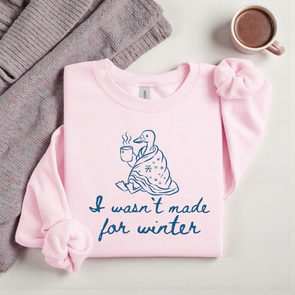Pink sweatshirt with a graphic and text, placed on a surface with a gray sweater and mug.